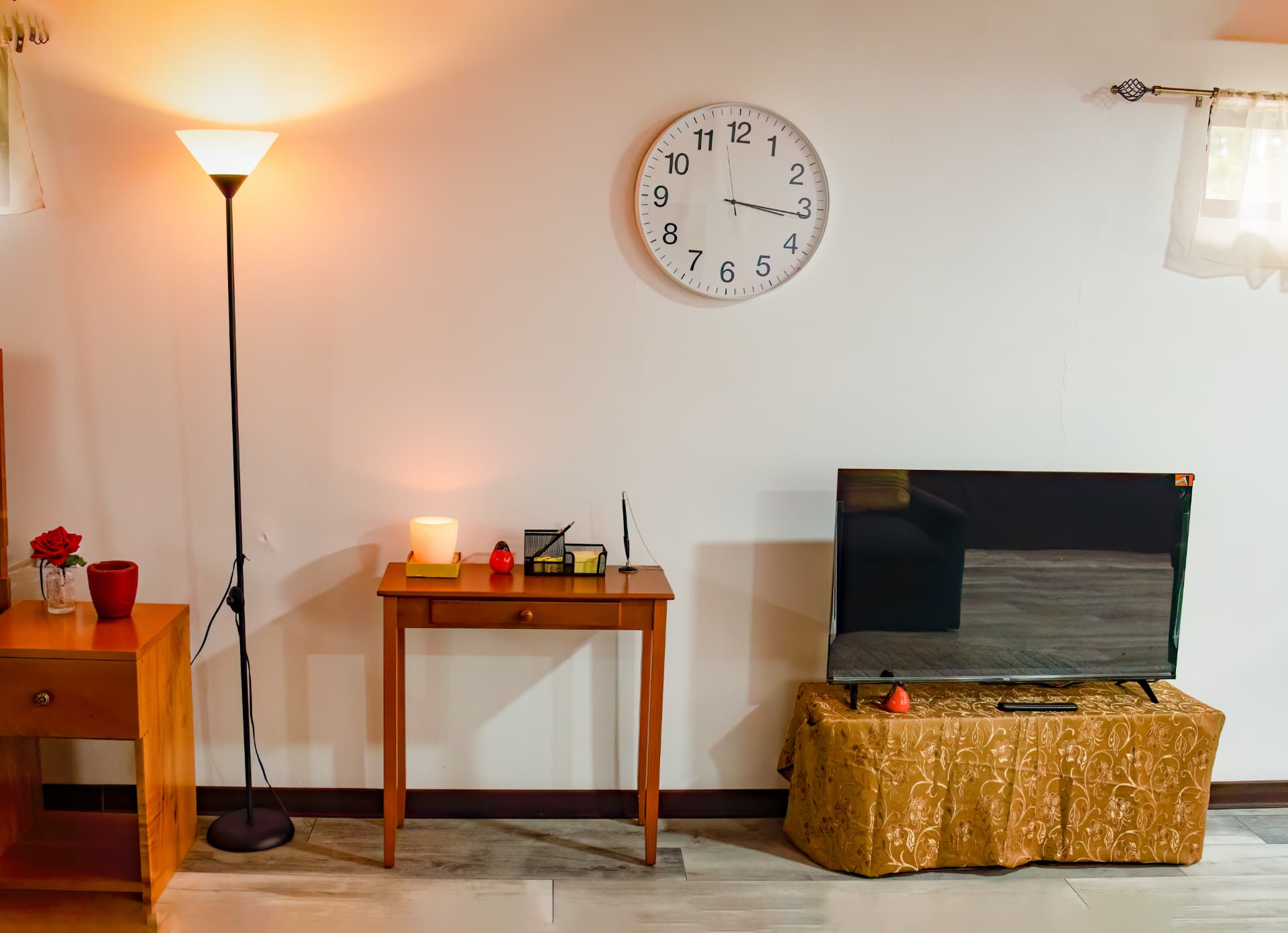 Smart TV on a draped table beside a writing table, floor lamp and wall clock